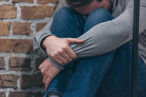 Vista recortada del hombre en suéter gris y pantalones azules sentado y abrazando las rodillas sobre fondo de textura marrón en la habitación, concepto de trastorno de duelo - foto de stock