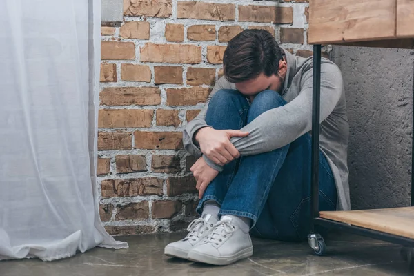 Hombre molesto sentado en el suelo en la esquina y abrazando las rodillas con la cabeza hacia abajo sobre fondo de textura marrón en la habitación, concepto de trastorno de duelo - foto de stock