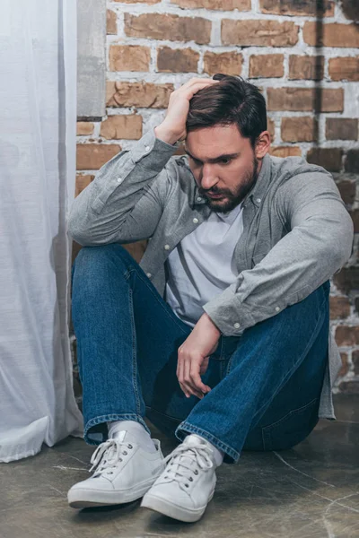 Upset man in grey shirt and blue pants sitting on floor on brown textured background in room, grieving disorder concept — Stock Photo