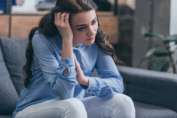 Sad woman in blue blouse sitting on grey couch and looking into distance at home, grieving disorder concept — Stock Photo