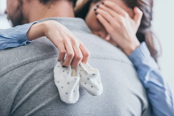 Visão cortada da mulher, abraçando o homem, segurando meias de bebê e chorando no quarto, conceito de transtorno de luto — Fotografia de Stock