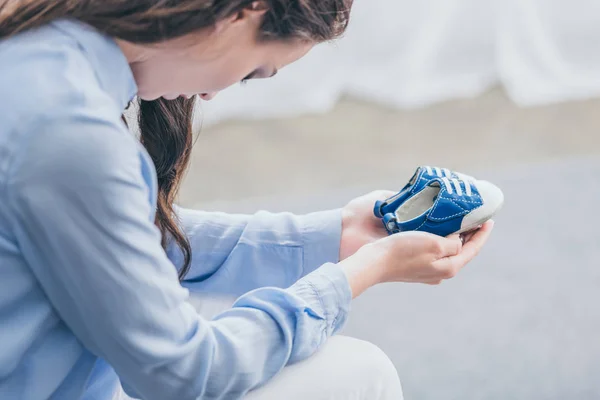 Cropped view of sad woman in blue blouse sitting and holding baby shoes at home, grieving disorder concept — Stock Photo