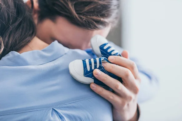 Visão cortada do homem abraçando a mulher e segurando sapatos de bebê azul no quarto, conceito de transtorno de luto — Fotografia de Stock