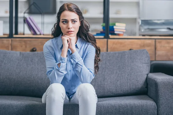 Sad woman in blue blouse and white pants sitting on grey couch in kitchengrieving disorder concept , — Stock Photo