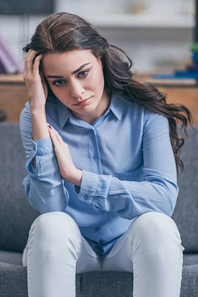 Sad woman in blue blouse and white pants sitting on grey couch at home, grieving disorder concept — Stock Photo