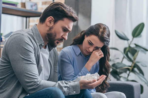 Woman sitting and crying on couch while man holding baby shoes in room, grieving disorder concept — Stock Photo
