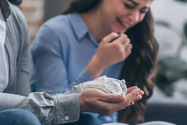 Vista recortada del hombre sosteniendo calcetines de bebé mientras que la mujer llorando en casa, concepto de trastorno de duelo - foto de stock