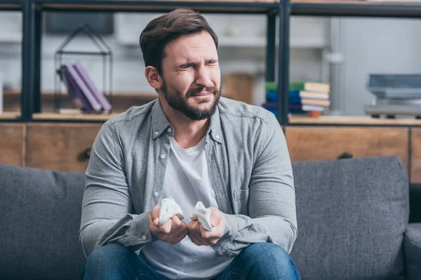 Homme en chemise grise et pantalon bleu assis sur le canapé, tenant des chaussettes de bébé et pleurant dans la chambre, concept de trouble du deuil — Photo de stock