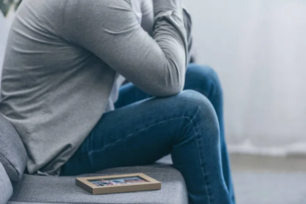 Vista ritagliata dell'uomo in camicia grigia e pantaloni blu seduti sul divano con cornice fotografica a casa, concetto di disturbo da lutto — Foto stock