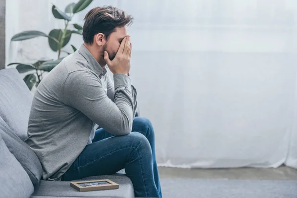 Hombre sentado en un sofá gris con foto en el marco y llorando en casa, concepto de trastorno de duelo - foto de stock