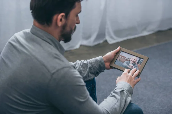 Cropped view of man sitting and looking at photo in frame with mature man at home, grieving disorder concept — Stock Photo