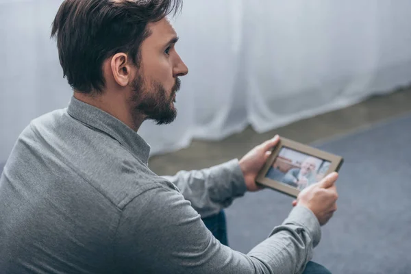 Ausgeschnittene Ansicht eines Mannes, der sitzt, Fotorahmen mit einem reifen Mann in der Hand hält und zu Hause in die Ferne blickt, Konzept der trauernden Unordnung — Stockfoto