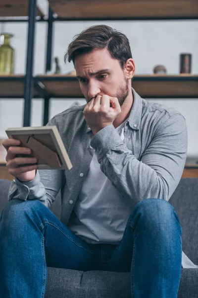 Trauriger Mann, der auf Couch sitzt und zu Hause ein Foto im Rahmen anschaut, trauert — Stockfoto