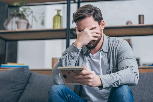 Upset man in gray shirt covering face with hand and holding photo frame at home, grieving disorder concept — Stock Photo