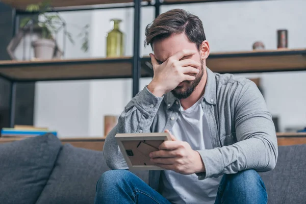 Hombre molesto en camisa gris cubriendo la cara con la mano y sosteniendo el marco de la foto en casa, concepto de trastorno de duelo - foto de stock