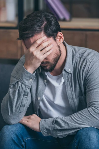 Расстроенный мужчина сидит, плачет и закрывает лицо рукой дома — стоковое фото