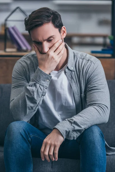 Hombre deprimido sentado, cubriendo la boca con la mano y afligiéndose en casa - foto de stock