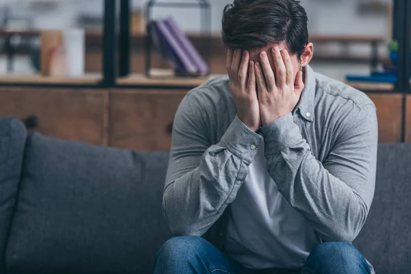 Hombre sentado en el sofá, llorando y y cubriendo la cara con las manos en casa - foto de stock