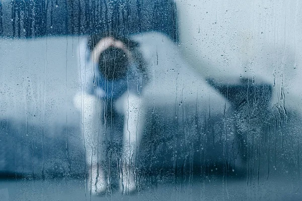 Подавленная женщина сидит на кровати и держит голову в руках через окно с каплей дождя — стоковое фото