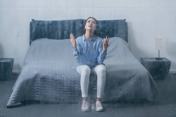 Mujer frustrada sentada en la cama, llorando y haciendo gestos con las manos en casa a través de la ventana con gotas de lluvia - foto de stock