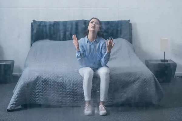 Femme frustrée assise sur le lit, pleurant et gesticulant avec les mains à la maison par la fenêtre avec des gouttes de pluie — Photo de stock