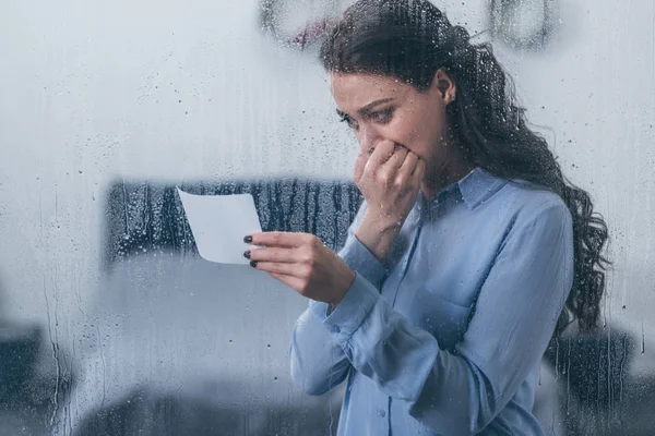 Trauernde Frau hält Foto in der Hand, bedeckt Mund mit Hand und weint zu Hause durch Fenster mit Regentropfen — Stockfoto