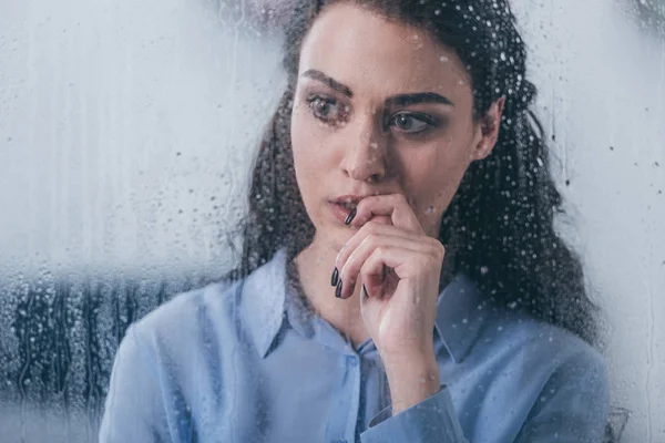 Красива засмучена жінка торкається обличчя і дивиться крізь вікно з краплями дощу — стокове фото
