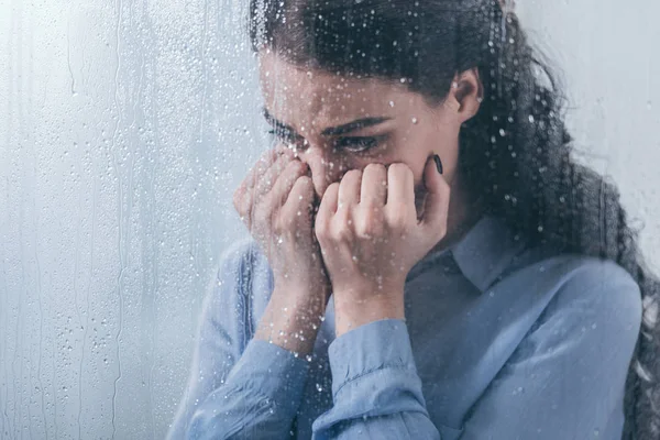Donna depressa che copre il viso con le mani e piange a casa attraverso la finestra con gocce di pioggia — Foto stock