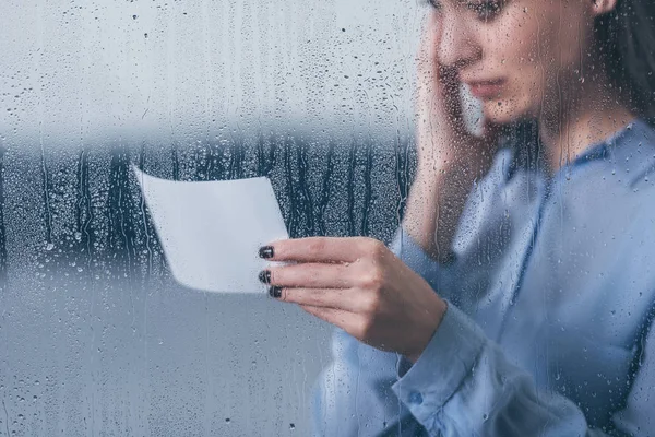 Vista recortada de la mujer afligida sosteniendo fotografía y llorando a través de la ventana con gotas de lluvia - foto de stock