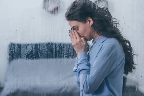 Depressed woman covering face with hands and crying at home through window with raindrops and copy space — Stock Photo