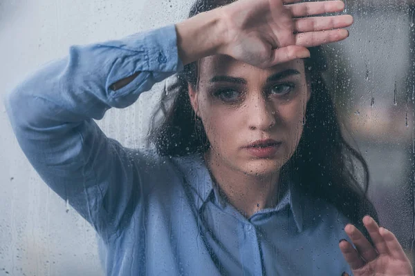Beautiful upset woman looking away and touching window with raindrops — Stock Photo