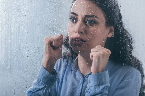 Belle femme triste avec les poings serrés regardant par la fenêtre avec des gouttes de pluie — Photo de stock