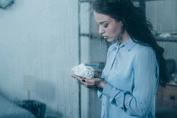 Grieving mother holding baby shoes through window with raindrops and copy space — Stock Photo