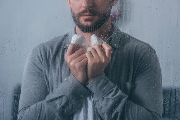 Cropped view of man holding baby shoes through window with raindrops — Stock Photo