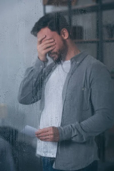 Depressed man covering face with hand, holding photograph and crying through window with raindrops — Stock Photo