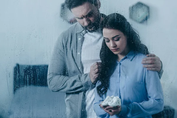 Mari deuil et étreinte femme avec des chaussures de bébé à travers la fenêtre avec des gouttes de pluie — Photo de stock