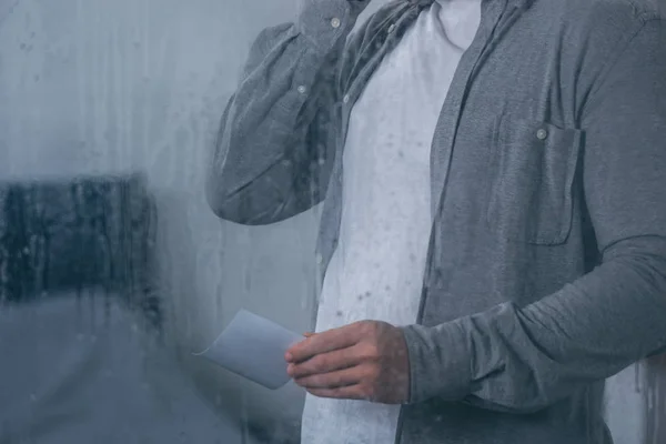 Cropped view of man holding photograph through window with raindrops — Stock Photo