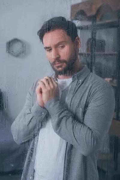 Upset man with folded hands through window with raindrops — Stock Photo