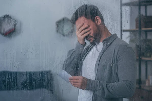 Depressed man covering face with hand, holding photograph and crying through window with raindrops — Stock Photo