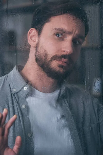 Handsome upset man touching window with raindrops and looking away — Stock Photo