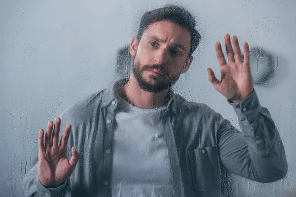 Bonito chateado homem tocando janela com gotas de chuva e olhando para longe — Fotografia de Stock