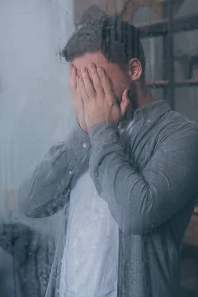 Adult man covering face with hands and crying through window with raindrops — Stock Photo