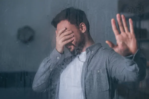 Hombre molesto cubriendo la cara con la mano, llorando y tocando la ventana con gotas de lluvia - foto de stock