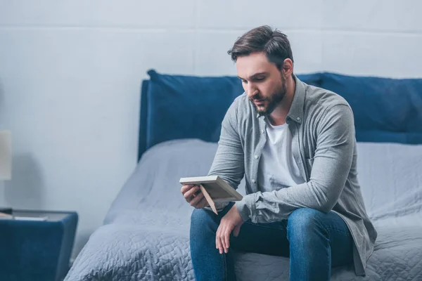 Grieving man sitting on bed and looking at photo frame at home with copy space — Stock Photo