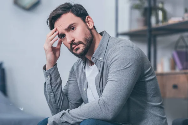 Selektiver Fokus eines gut aussehenden, aufgebrachten Mannes, der sitzt, das Gesicht berührt und zu Hause trauert — Stockfoto