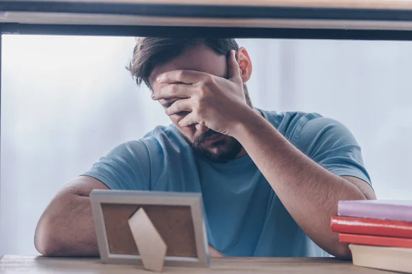 Hombre cubriendo la cara con la mano y el duelo cerca del marco de fotos en casa - foto de stock