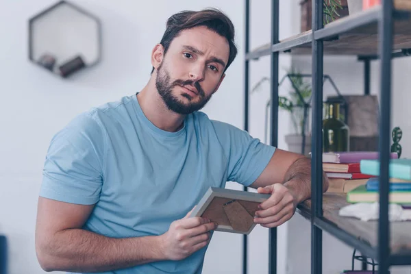 Selective focus of upset man holding photo frame and looking at camera at home — Stock Photo