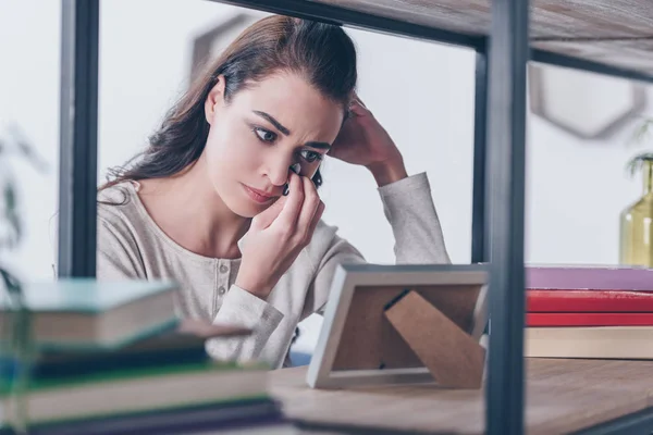 Selektiver Fokus einer aufgebrachten Frau, die auf Bilderrahmen blickt, zu Hause weint und Tränen wischt — Stockfoto