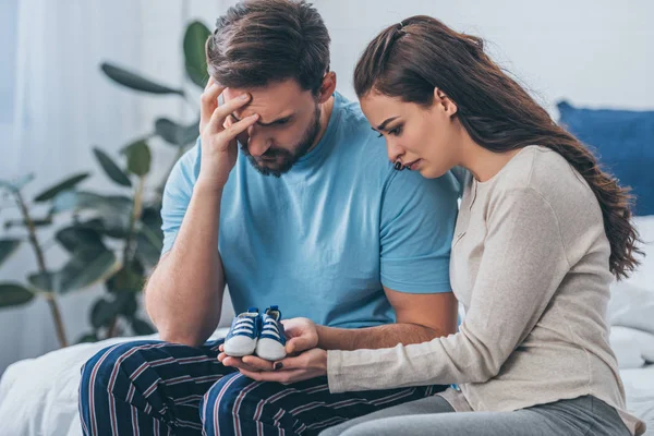 Selektiver Fokus trauernder Eltern, die auf dem Bett sitzen und Babyschuhe zu Hause halten — Stockfoto