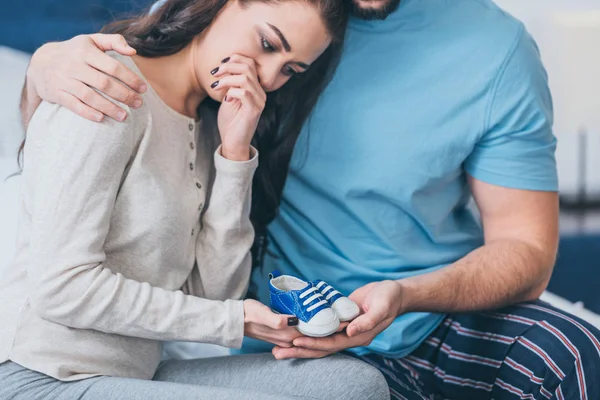 Selective focus of husband hugging grieving and crying wife with baby shoes — Stock Photo